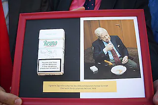 FOTOS 1699, 1700: IRENE BÄR<?ZE?>DSCN 1699 Übergabe von Helmut Schmidts Zigarrettenschachtel, von links: <?ZE?>Karl-Rainer Kopf (stellvertretender SPD-Kreisvorsitzender, Ortenau), MdB, SPD Johannes Fechner, Bürgermeister Dietmar Benz, Museumsleiter Patrick Benz, Fördervereinsvorsitzender Wolfgang Ohnemus DSCN 1700 Helmut Schmidts handsignierte Zigarrettenschachtel<?ZE?><?ZE?>MAHLBERG (ib). Manch verdientem staatstragendem Politiker ergeht es wie Rockstars zu Lebzeiten: Dinge des täglichen Gebrauchs, die sie einmal in ihren Händen hatten, werden von anderen als begeherte Trophäe begehrt. <?ZE?>In dem Fall ist es eine Zigarrettenschachtel von Altbundeskanzler Helmut Schmidt, die in der Sonderausstellung des Tabakmuseums ihr würdiges Plätzlein finden wird. Ergattert hatte sie MdB Johannes Fechner von der SPD, der sie am Dienstag Nachmittag Vertretern des Tabakmuseums, der Stadt und des Fördervereins übergab.<?ZE?>Nicht nur für Fechner, dem der passionierte Kettenraucher Schmidt seine leere Schachtel mit Mentholzigarretten der Marke Reyno mit einem Autogramm versehen gab, war's das Highlight des Tages. Sichtlich stolz präsentierte der Nichtraucher die Schachtel auf einem Rahmen neben einem Bild, das Helmut Schmidt in Aktion beziehungsweise beim Rauchen zeigt; mit der Übergabe hat er sein Versprechen eingelöst, das er im Juni beim Museumsfest gemacht hat. "Für uns ist es etwas Besonderes, wenn eine solche Devonotialie überbracht wird", sagte der stellvertretende Kreisvorvorsitzende Karl-Rainer Kopf. Eine große Freude war's auch für Bürgermeister Dietmar Benz, Museumsleiter Patrick Benz und Wolfgang Ohnemus als Vorsitzender des Fördervereins fürs Tabakmuseum. "Mir ist ein rauchender Helmut Schmidt lieber als ein nichtrauchender Politiker ohne Format", so Ohnemus. Als Mitglied der CDU sprach er mit "größter Hochachtung, Anerkennung und Bewunderung" von einem Bundeskanzler, der viele seiner Vorgänger in den Schatten gestellt habe und an dem sich viele Nachfolger ein Beispiel nehmen sollten.<?ZE?>Strikt überzeugte Nichtraucher stören sich sich daran, wenn Schmidt im Fernsehen bei Talkshows und anderen öffentlichen Auftritten raucht. <?ZE?>Sei's drum, laut Museumsleiter Patrick Benz hat sich der "letzte öffentlich-rechtliche Raucher" mit seiner abgelegten Zigarrettenschachtel einen Platz neben den Zigarren von Ludwig Erhardt, Gerhardt Schröder, Fidel Castro und Schwedens König Gustav VI sowie einer Pfeife von Herbert Wehner im Oberrheinischen Tabakmuseum verdient gemacht. - Natürlich weniger um des Rauchens als um seiner Verdienste willen. Dass nebenbei gesagt im Fernsehen eigentlich keine Pfeifen, Zigarren oder Zigarretten mehr zu sehen seien, sei positiv zu sehen, weil dadurch die Vorbildfunktion des Rauchens entfalle. Nichts desto weniger war der Museumsleiter im Rückblick auf die frühere Kultur des Rauchens davon beeindruckt, wie Teilnehmer des Internationalen Früschoppens mit Werner Höfer globale Probleme wälzten und dabei das Fernsehstudio "so richtig einqualmten". Aber irgendwie gehören Politik und Rauchen wohl immer noch zusammen, wenn man Helmut Schmidt glauben darf. Benz zitierte seinen Vergleich von der Politik mit dem Kampfsport, in dem vor allem zwei Dinge vonnöten seien. "Willen braucht man und Zigaretten."<?ZE?>Vom Glanz des Altbundeskanzlers respektive des Abglanzes seiner Zigarrettenschachtel hoffte seinerseits Dietmar Benz, dass da auch ein bisschen davon auf das Tabakmuseum abfällt. "Es wird mit der Spende aufgewertet." Das Museum, in dem seit auf mehr als 1700 Quadratmetern mit dem Thema Tabak ein Teil der früheren regionalen Wirtschaftsgeschichte gezeigt wird, kann mehr Besucher gut gebrauchen. <?ZE?>Der Bürgermeister erinnerte auch an den Kauf der ehemaligen Fabrikantenvilla vor gut zwei Jahren; zum Beginn der neuen Museumssaison soll die wieder mit dem Museum und anderen Nebegebäuden als optisches Ensemble wieder hergestellt sein. Bis dahin wird Helmut Schmidt, der sich laut Fechner übrigens "sehr interessiert" am Mahlberger Kleinod gezeigt habe, sein Dankeschön erhalten haben, und zwar einen Film über die Kultur- und Wirtschaftsgeschichte des Tabaks in der hiesigen Region. [Irene Bär]